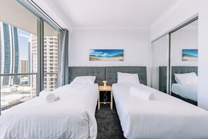 Habitación de hotel con dos camas, una mesita de noche, una lámpara y un ventanal con vistas a la ciudad.