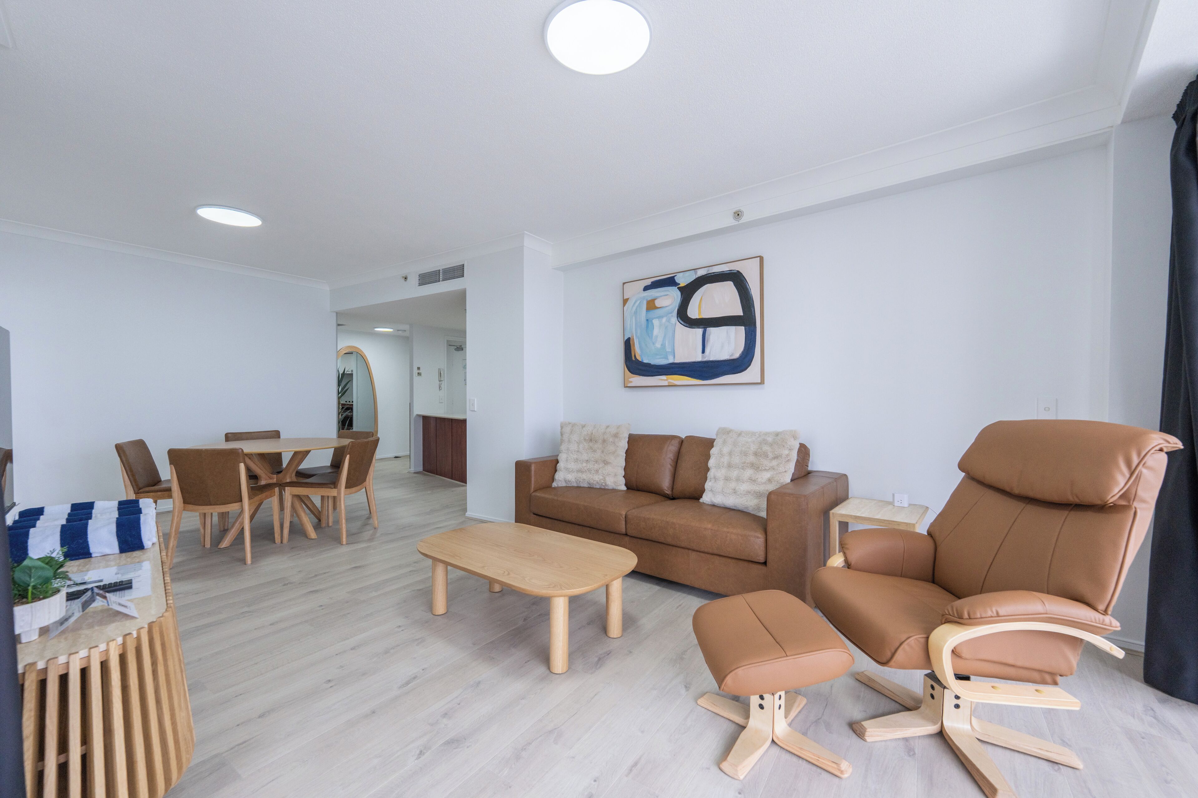 A modern living room with a brown leather sofa, wooden coffee table, and matching ottoman. There is a dining area with a wooden table and chairs, and a painting on the wall.