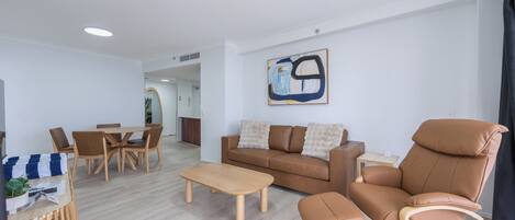 A modern living room with a brown leather sofa, wooden coffee table, and matching ottoman. There is a dining area with a wooden table and chairs, and a painting on the wall.