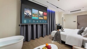 Une chambre d’hôtel avec un lit, un écran de télévision affichant une sélection de films, une baignoire et un panier de fruits sur la table de chevet.
