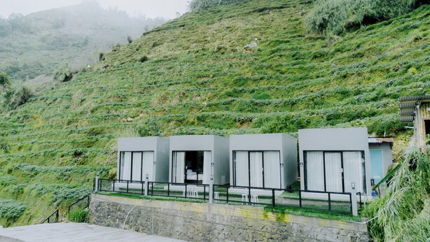 A hillside with a row of modern, two-story buildings with large windows and a stone retaining wall in the foreground. - Stigma Kubus By Travelink (Kejajar)