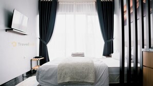 A modern hotel room with a bed, a flat-screen TV mounted on the wall, a small wooden side table, and a window with dark curtains. - Stigma Kubus By Travelink (Kejajar)