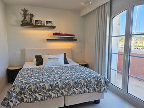 Un dormitorio con cama, mesitas de noche, una ventana y una estantería con objetos decorativos.
