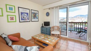 A living room with a sofa, a coffee table, and a TV. There are framed pictures on the wall and a balcony with a view of the city.