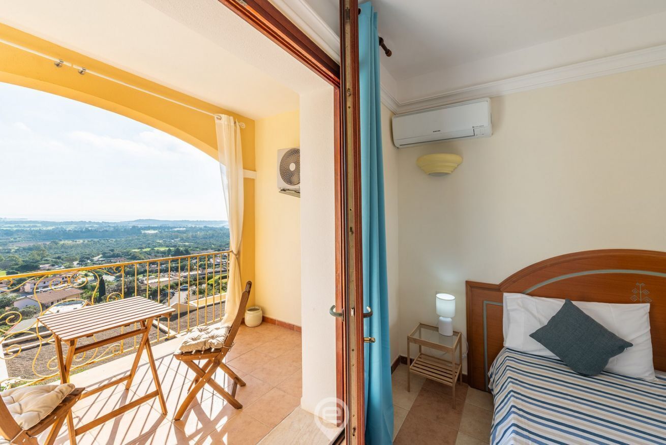 Un balcón con una mesa y sillas de madera, una cama con colcha a rayas y vistas al paisaje.