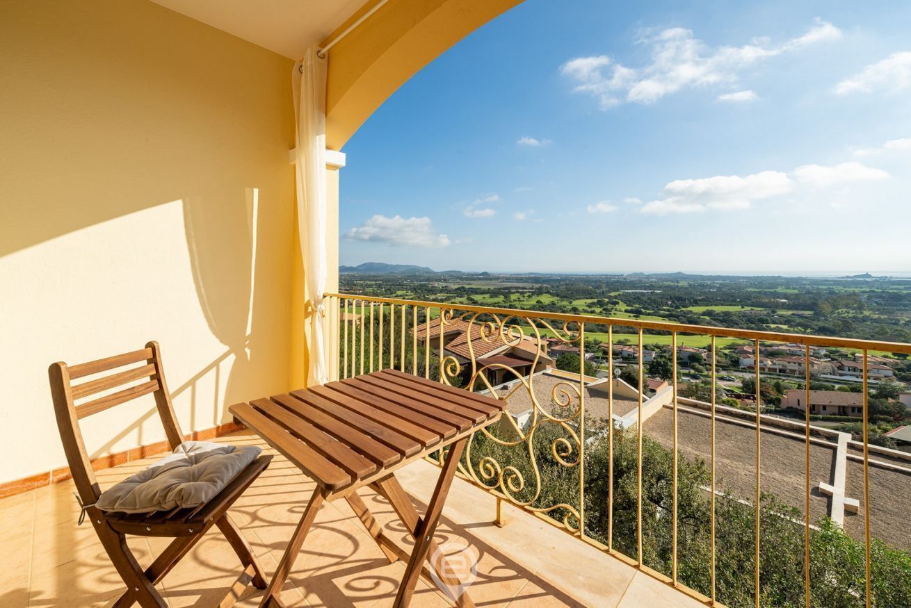 A balcony with a wooden table and chair, a yellow wall, and a view of a residential area with houses and greenery.