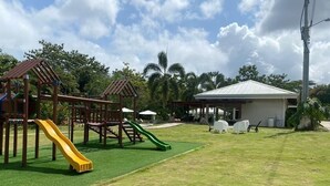 Ein Spielplatz mit Rutschen und Klettergerüsten, einem überdachten Sitzbereich und einem Gebäude mit Satteldach.