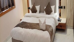 Una cama bien hecha con una almohada y una toalla doblada, una mesita de noche con un teléfono, y una lámpara fijada a la pared.