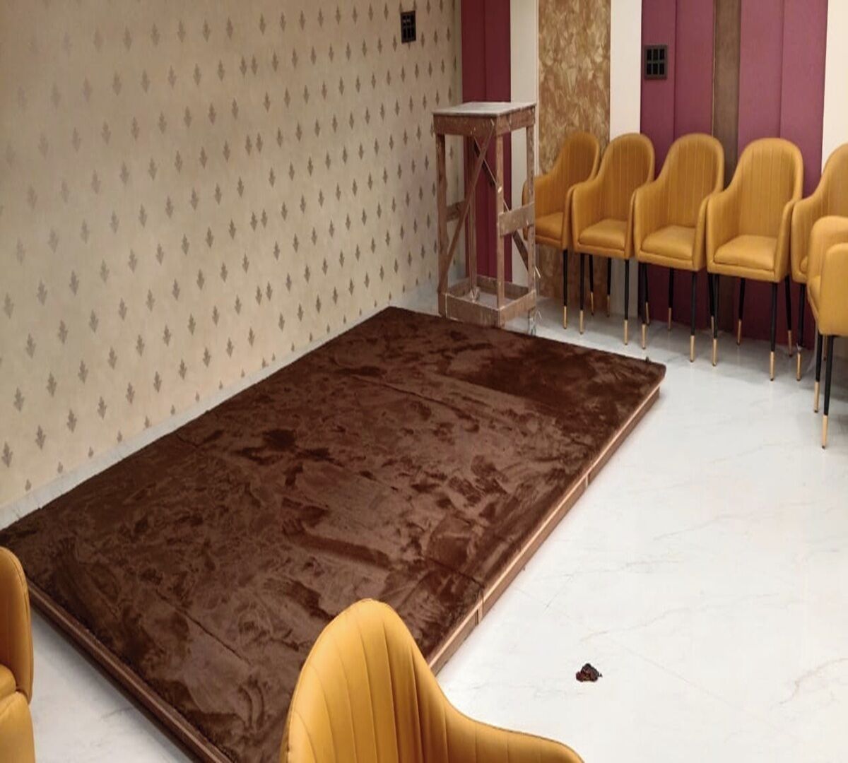 A room with a large brown carpet, a small table, and several yellow chairs.