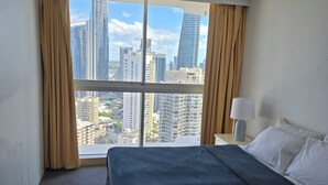 Ein Hotelzimmer mit Bett, Deckenventilator, Nachttisch mit Lampe und großem Fenster mit Blick auf die Stadt.