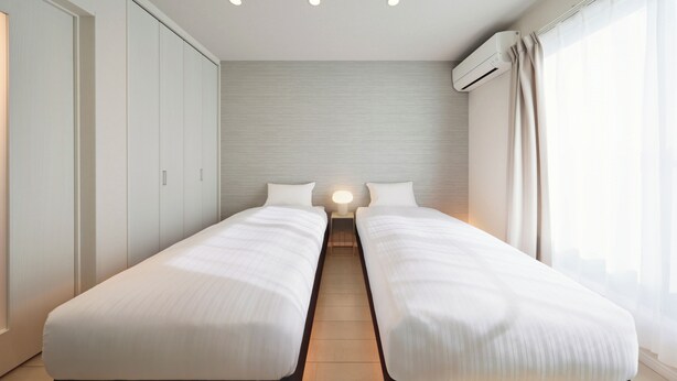 A modern hotel room with two single beds, a nightstand with a lamp, a wardrobe, and an air conditioning unit. - Crystalcourt Shirahama (Shirahama)