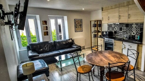 Una sala moderna con un sofá de cuero negro, una mesa de centro de vidrio y una mesa de comedor de madera con sillas. Se aprecia una cocina al fondo, con un salpicadero de diseño y armarios de madera.