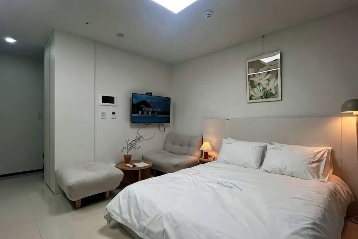 A hotel room with a bed, a chair, a small table, a TV, and a wall-mounted lamp.