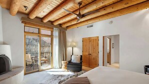 A bedroom with a large bed, wooden ceiling beams, a fireplace, and a sliding glass door leading to an outdoor area with a chair and table. - Incredible Sunset Views, Community Pool (Santa Fe)