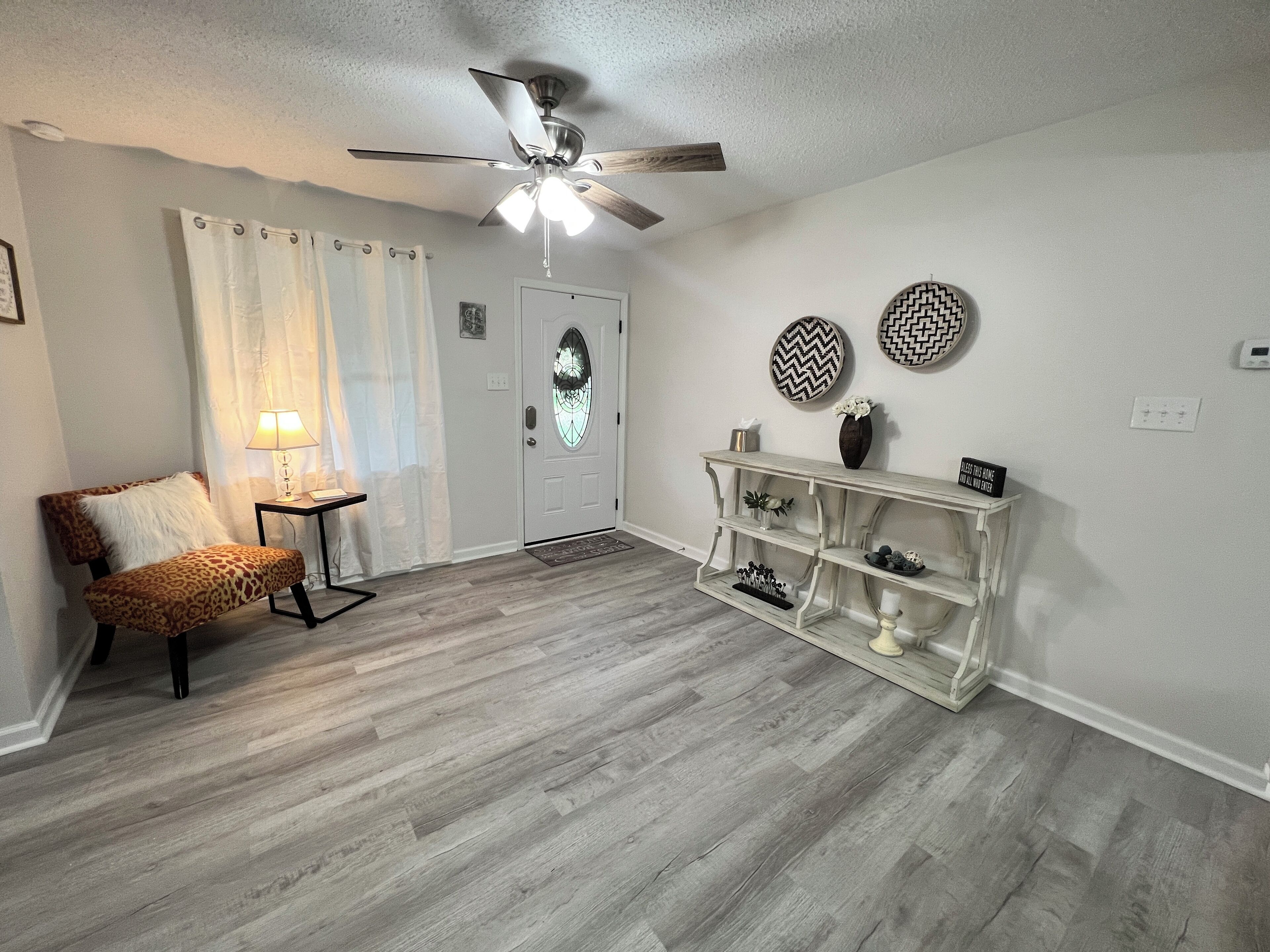 Una sala de estar con ventilador de techo, una puerta blanca con panel de vidrio decorativo, un sillón estampado de leopardo, una mesita con lámpara y una consola blanca con platos decorativos.