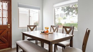 Une salle à manger avec une table en bois et quatre chaises, une porte en bois et deux fenêtres équipées de stores.