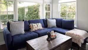 Un canapé bleu agrémenté de coussins à rayures et à motifs, une table basse en bois ornée d’objets décoratifs et de grandes fenêtres offrant une vue sur la verdure.