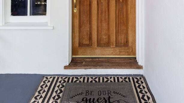 Une porte d’entrée en bois ornée d’une guirlande, avec une boîte aux lettres et un paillasson sur lequel est inscrit : « SOYEZ LES BIENVENUS ».