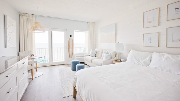 Une chambre avec un grand lit, une commode blanche, une table ronde et un balcon où se trouve une planche de surf.