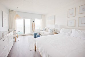 A bedroom with a large bed, a white dresser, a round table, and a balcony with a surfboard. - Hampton Tides | Ocean Views | Top Floor Balcony (Hampton)