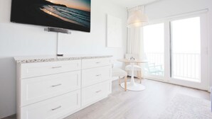 Une chambre d’hôtel moderne dotée d’un grand téléviseur à écran plat fixé au-dessus d’une commode blanche, d’une table ronde avec des chaises et d’un balcon offrant une vue sur l’océan.