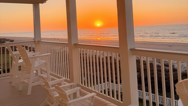 A wooden deck with white chairs facing a sunset over the ocean.