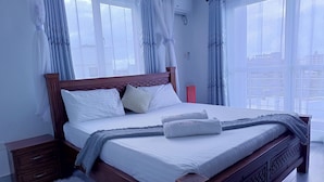 Una cama bien hecha con sábanas blancas y una manta gris, cabecera de madera y mesita de noche con lámpara.