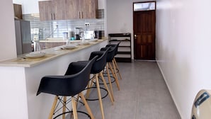 Una cocina moderna con armarios de madera, una encimera y taburetes de bar. Hay una puerta que da a otra habitación.