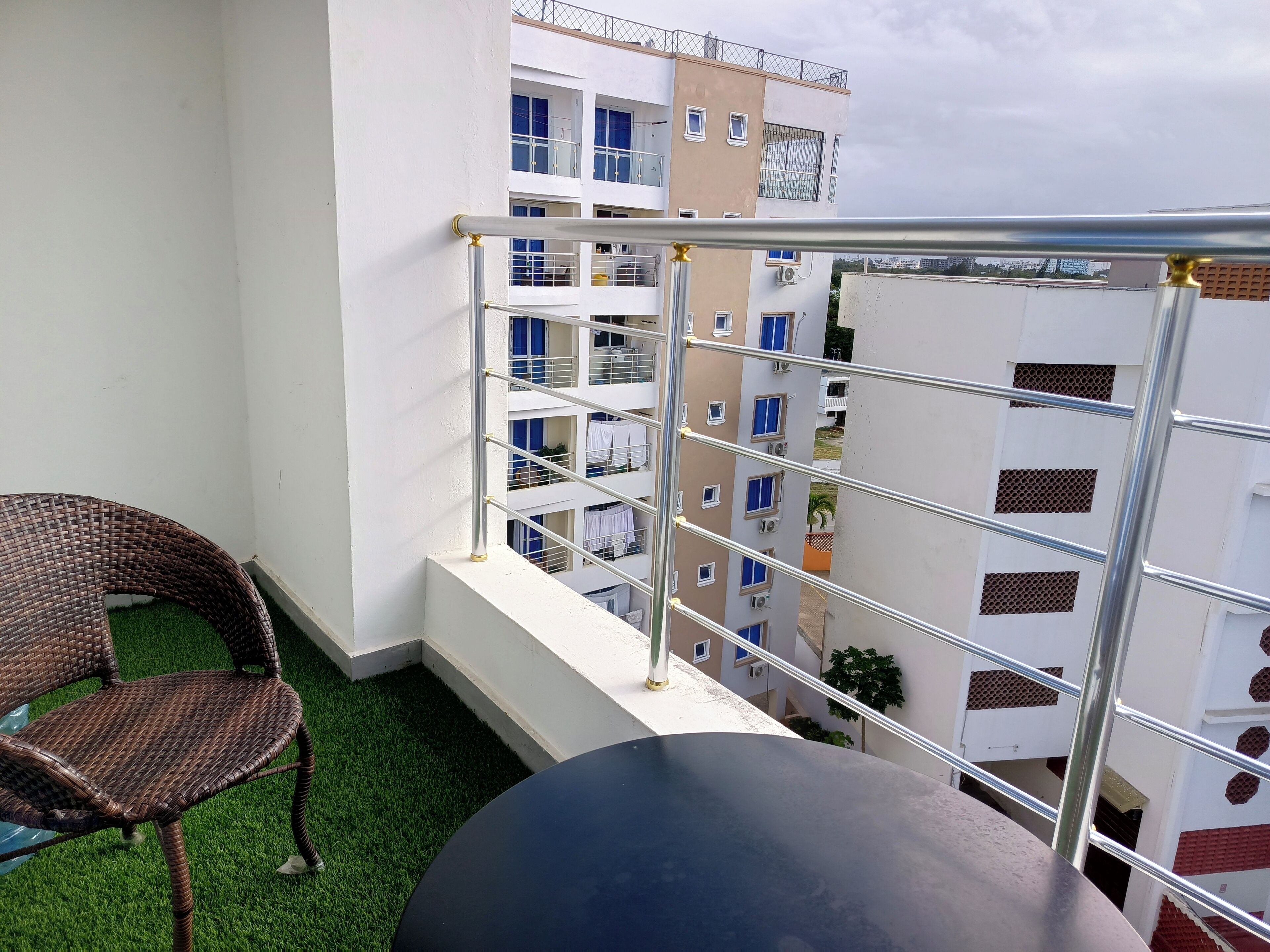 Un balcón con una silla de mimbre y una mesa redonda, con vista a un edificio de varios pisos con balcones y unidades de aire acondicionado.