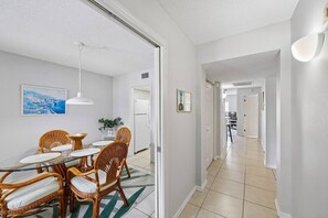 A dining area with wicker chairs and a glass table, a hallway with a tiled floor, and a view into another room with a TV.