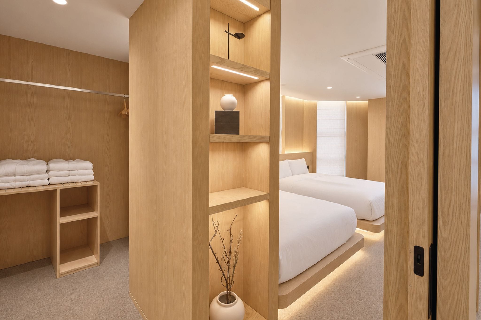 A modern hotel room with wooden paneling, a bed, a bedside table, a wardrobe, and a shelf unit with a vase.