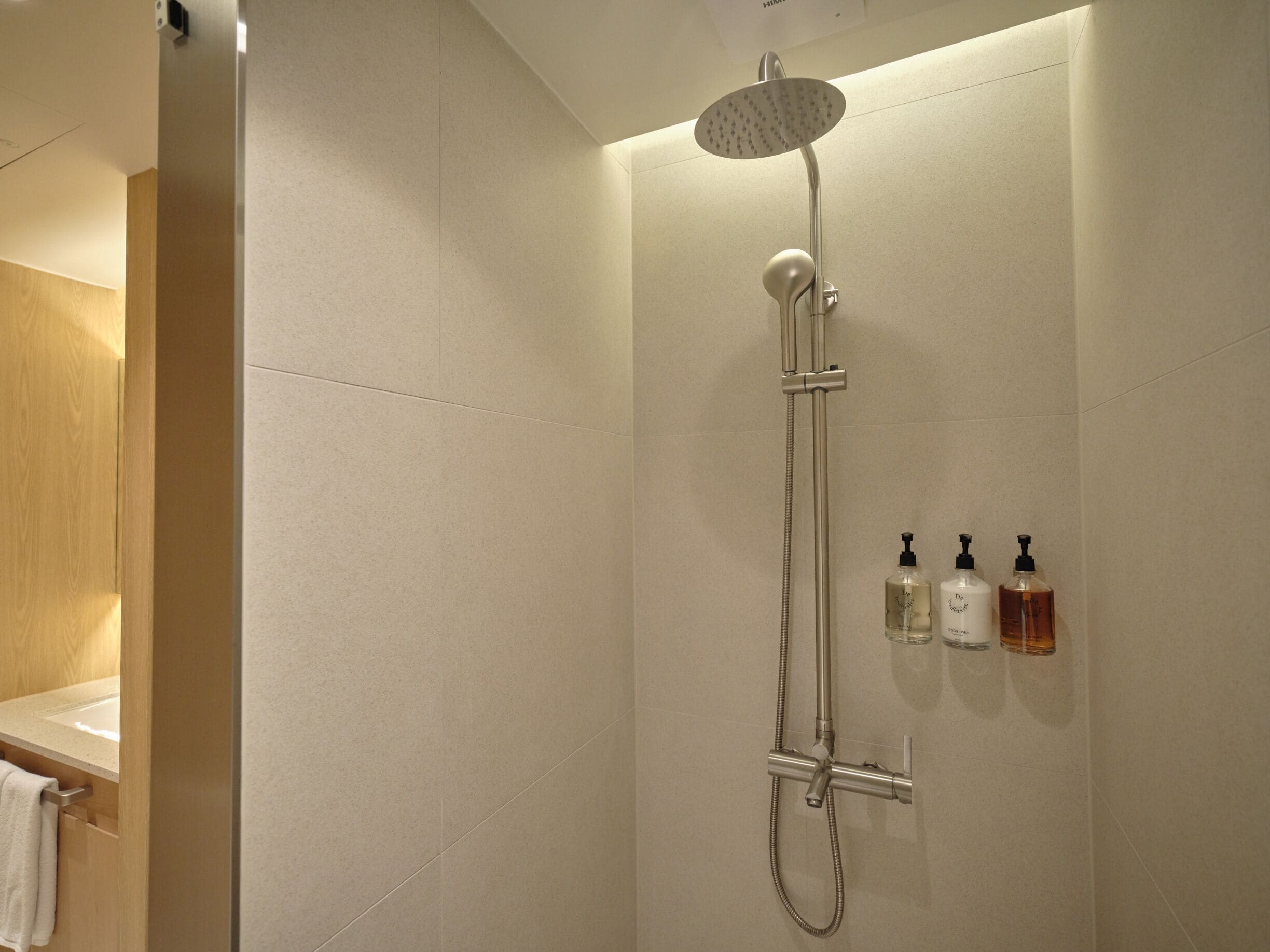 A shower with a handheld showerhead and a handheld hose, a towel rack, and three bottles of shampoo and conditioner on the wall.