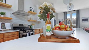 Una cocina con encimera blanca, una botella de vino, un bol de manzanas y un jarrón con flores.