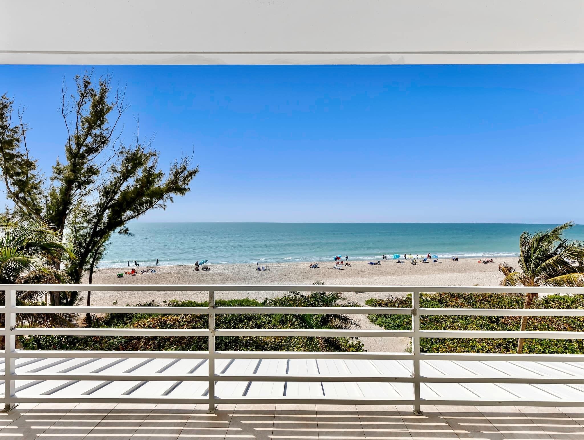 A beach view from a balcony with a railing, palm trees, and a clear blue sky.