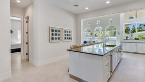 A modern kitchen with a central island, white cabinetry, and large windows offering views of the outdoors. - Casa Captiva - Ground Level 5 BR Luxe Villa with Grotto Pool, Steps to Beach (Captiva)
