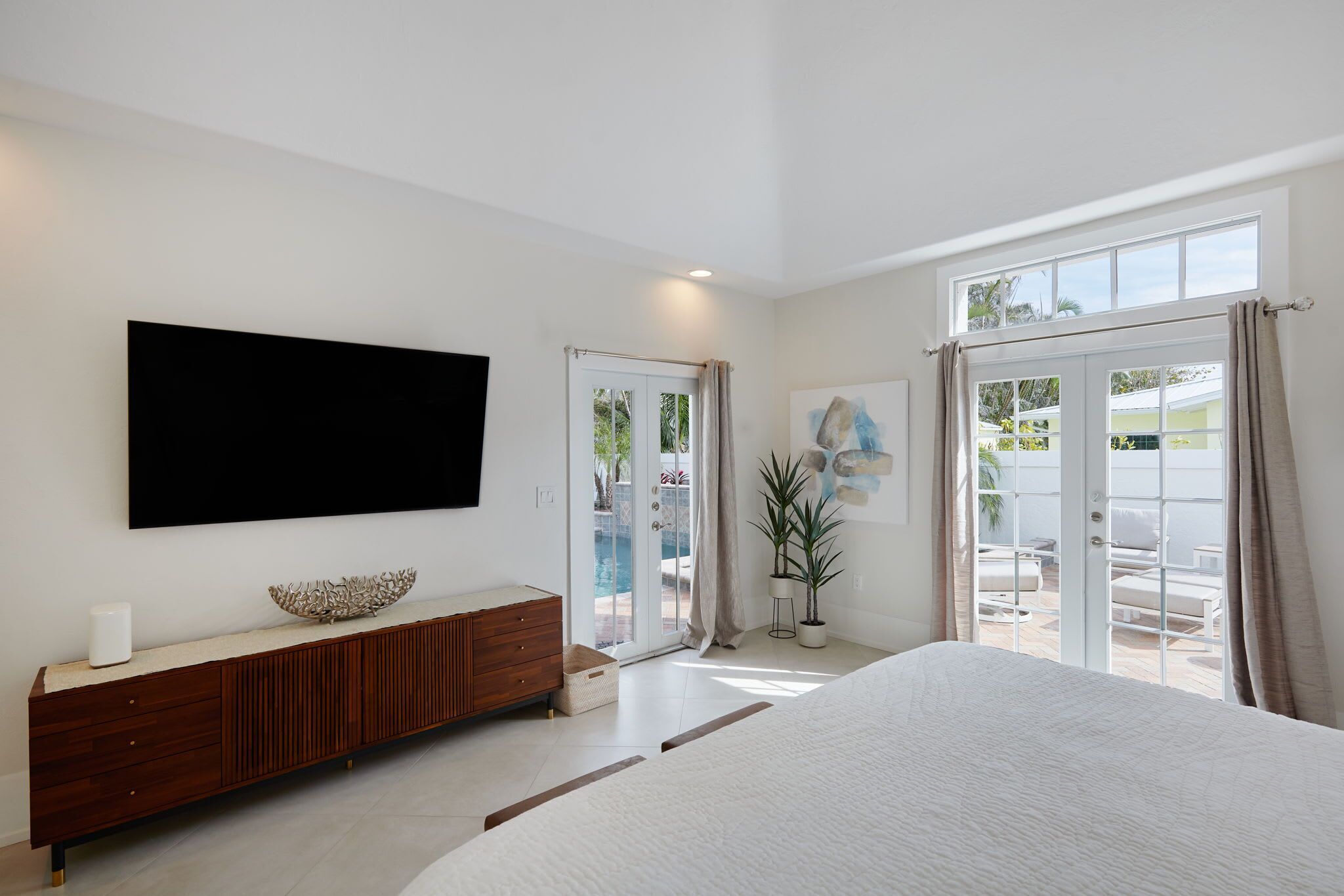 A modern bedroom with a large bed, a TV mounted on the wall, a wooden dresser, and a sliding glass door leading to an outdoor area.
