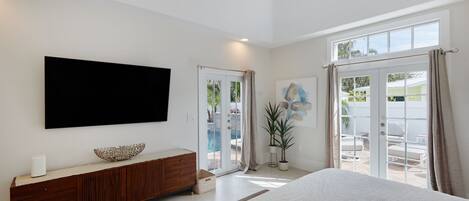A modern bedroom with a large bed, a TV mounted on the wall, a wooden dresser, and a sliding glass door leading to an outdoor area.