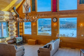 A log cabin interior with a fireplace, two sofas, large windows, and a chandelier. - FOXFHILL: 6,000+ sq ft log estate, 4BR/5BA sleeps 16. (Otis Orchards)