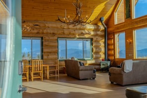 A log cabin interior with a dining area, two sofas, a fireplace, and large windows offering mountain views. - FOXFHILL: 6,000+ sq ft log estate, 4BR/5BA sleeps 16. (Otis Orchards)