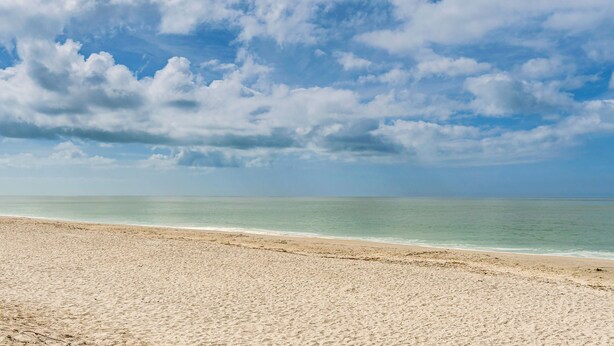 Une plage dotée d’un large rivage sablonneux et offrant une vue dégagée sur l’océan, sous un ciel partiellement nuageux.