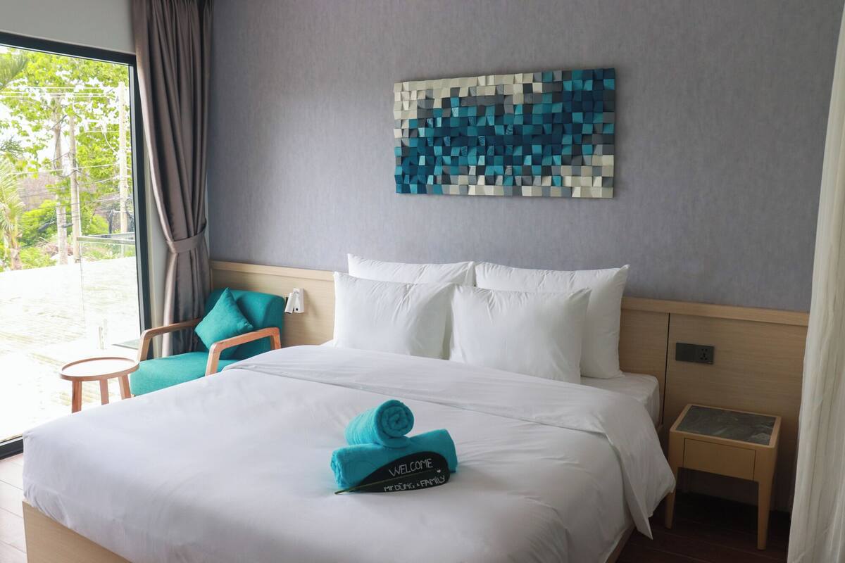 Una habitación de hotel con cama, una silla color turquesa, una mesita de noche y una obra de arte en la pared.