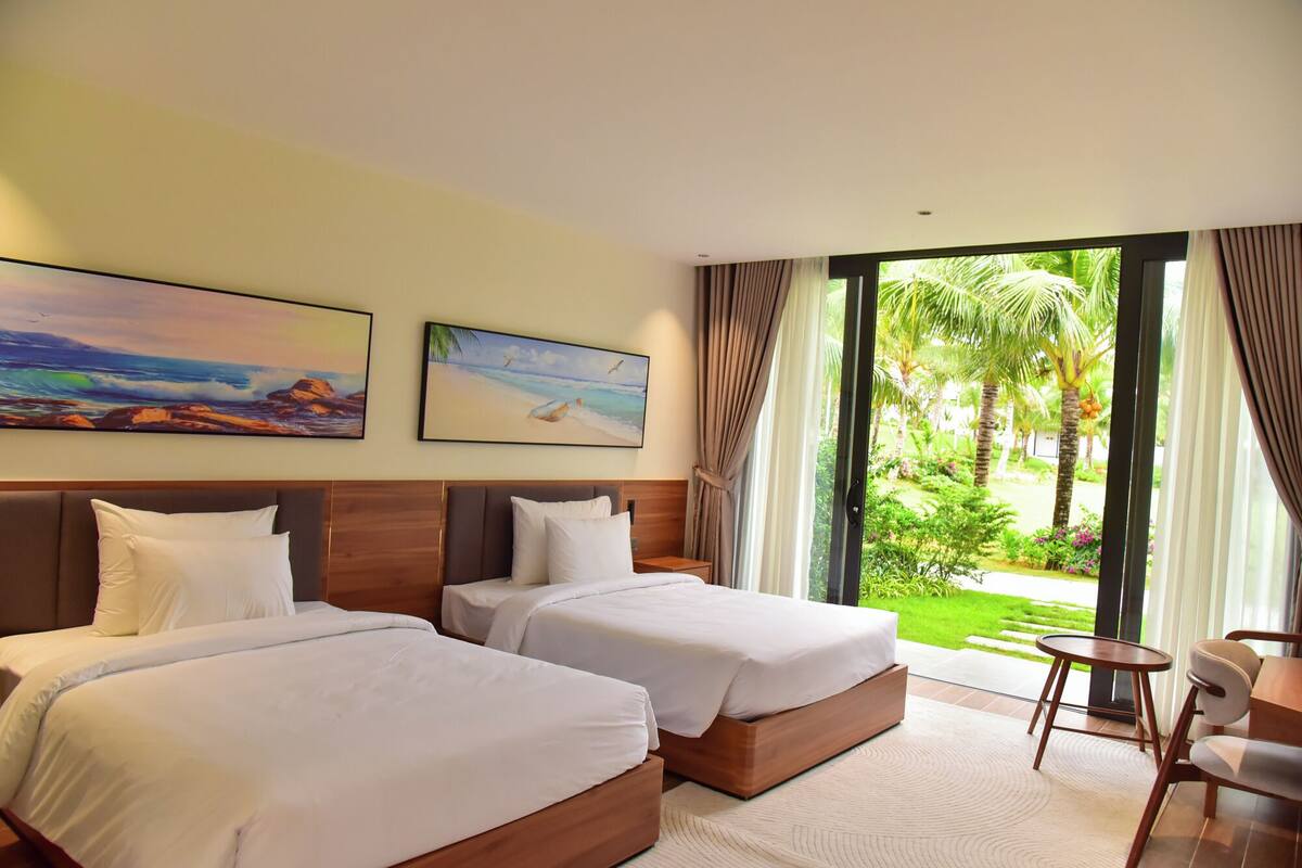 Habitación de hotel con dos camas, una mesita y una silla. Hay dos cuadros en la pared y una vista a un jardín desde la ventana.