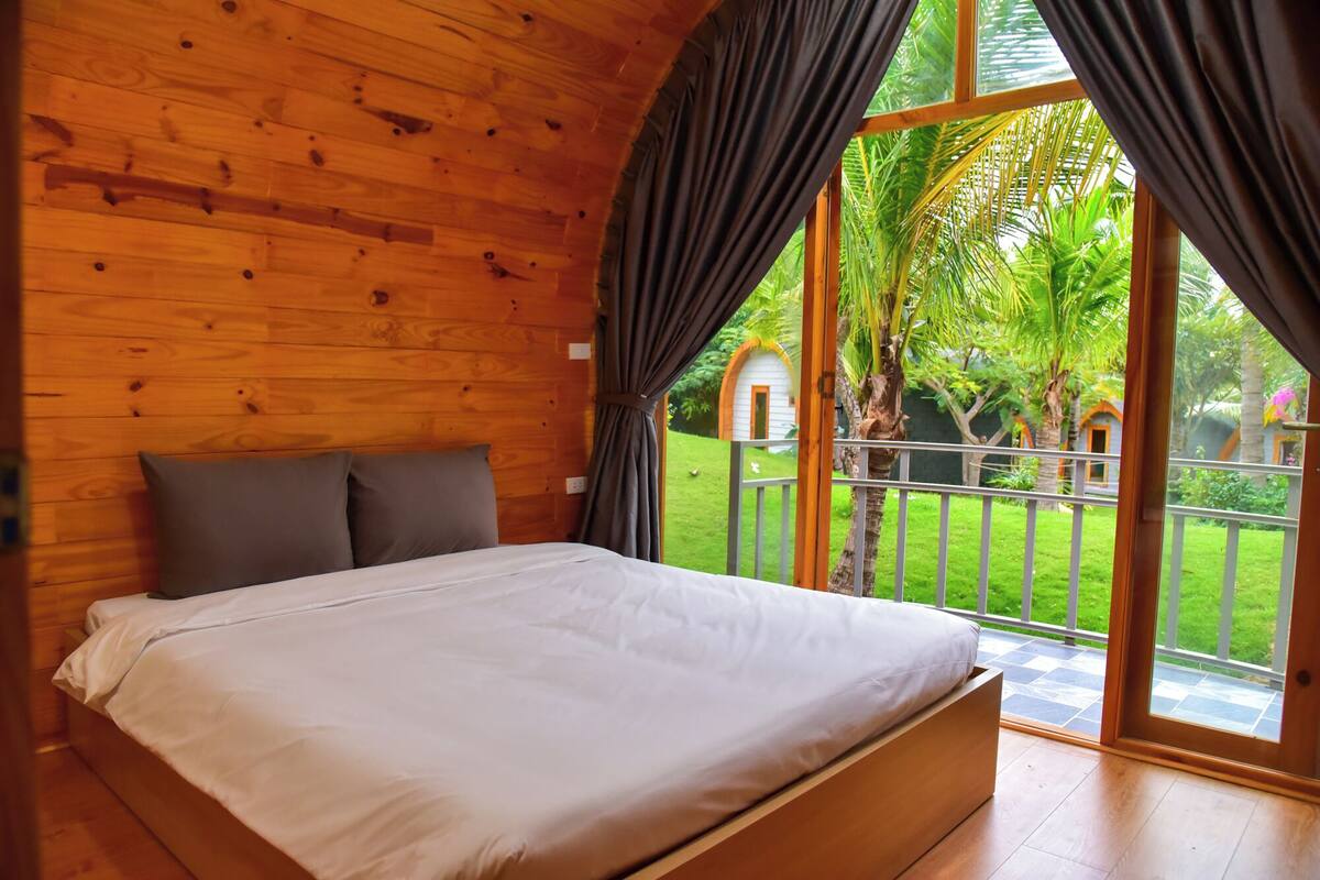 Un dormitorio con paneles de madera, una cama, cortinas grises y vistas a un jardín a través de amplios ventanales.