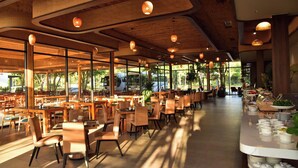A spacious restaurant with long tables and chairs, large windows, and hanging pendant lights.