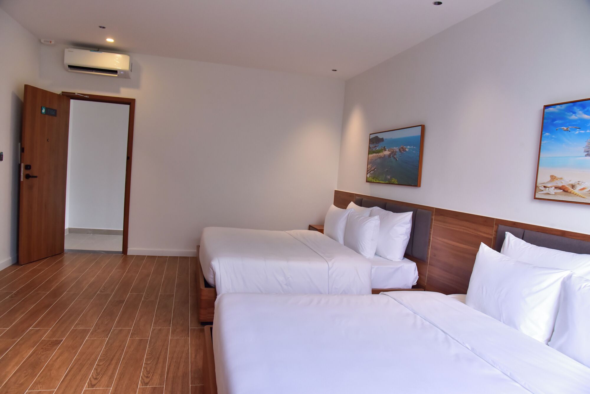 A hotel room with two beds, a wooden floor, a painting of a beach, and a door.