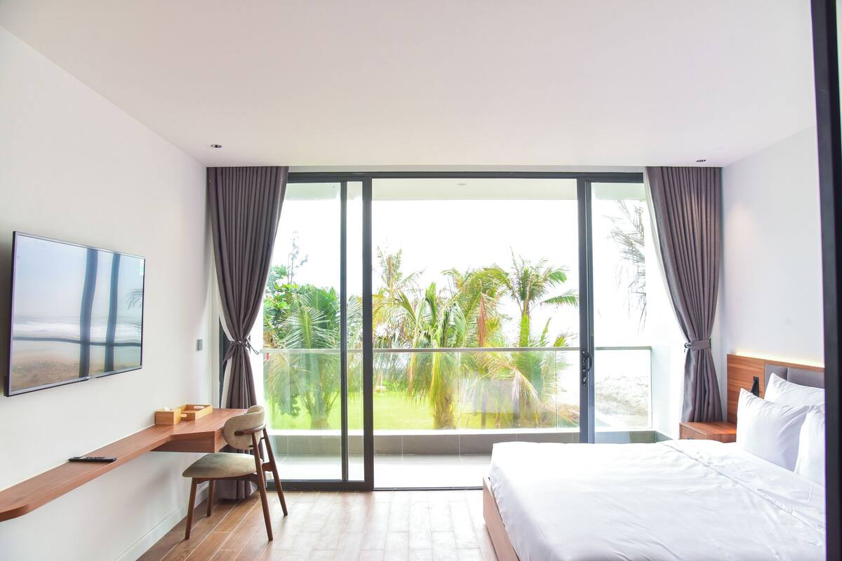 Una habitación de hotel con una cama, un escritorio con una silla, un televisor montado en la pared y un balcón con vistas a palmeras.