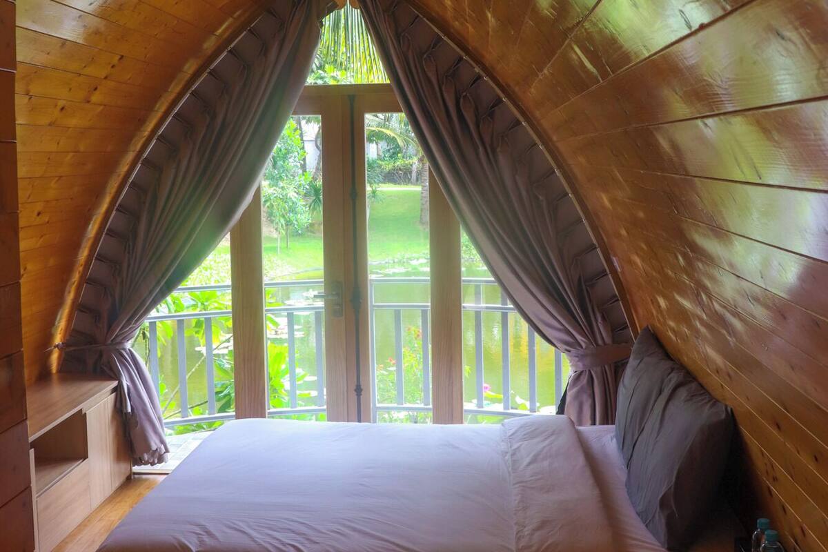 Un dormitorio con techo inclinado de madera, una cama, una mesita de noche y vistas a un paisaje verde a través de una ventana.