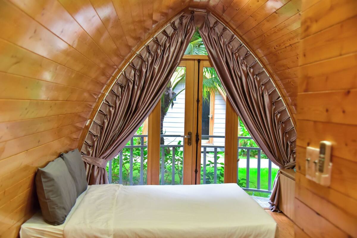 Acogedor dormitorio en ático con cama, paredes de madera y vista a la vegetación exterior.