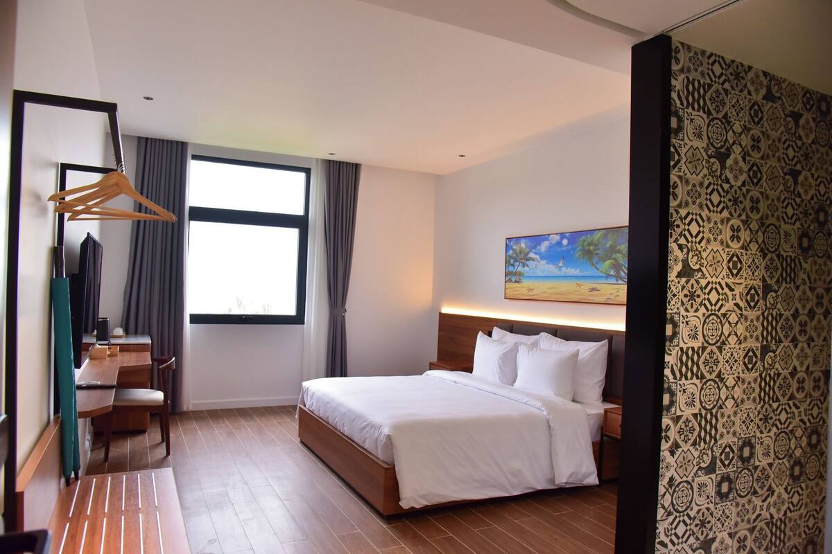 Una habitación de hotel con una cama, un escritorio con una silla, una ventana y un cuadro en la pared.