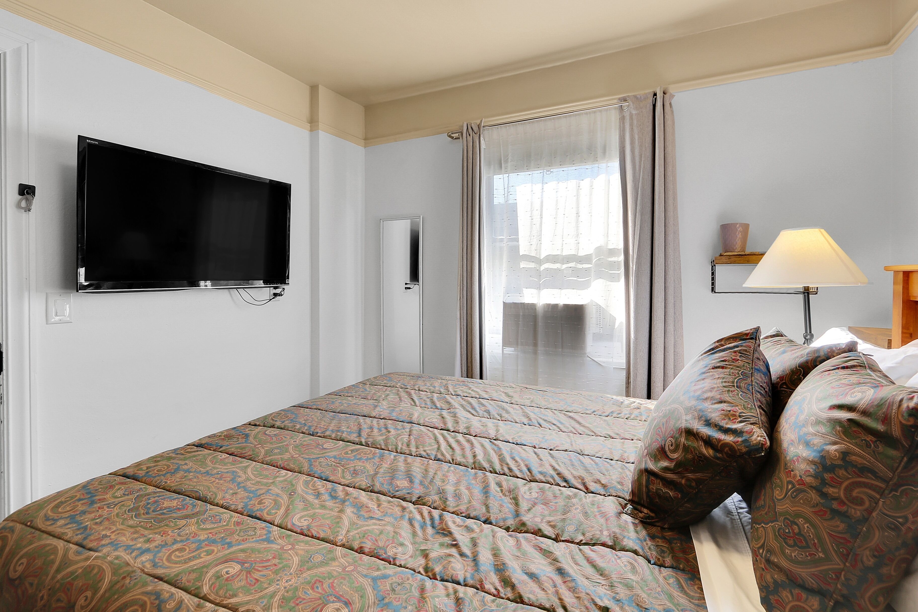 Hotelkamer met een bed, een aan de muur bevestigde televisie, een raam met gordijnen en een nachtkastje met een lamp.